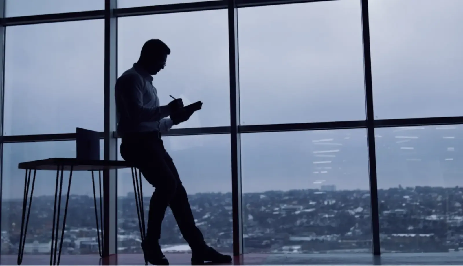 Silhouette of a businessman writing in a notebook beside a high-rise office window overlooking a city